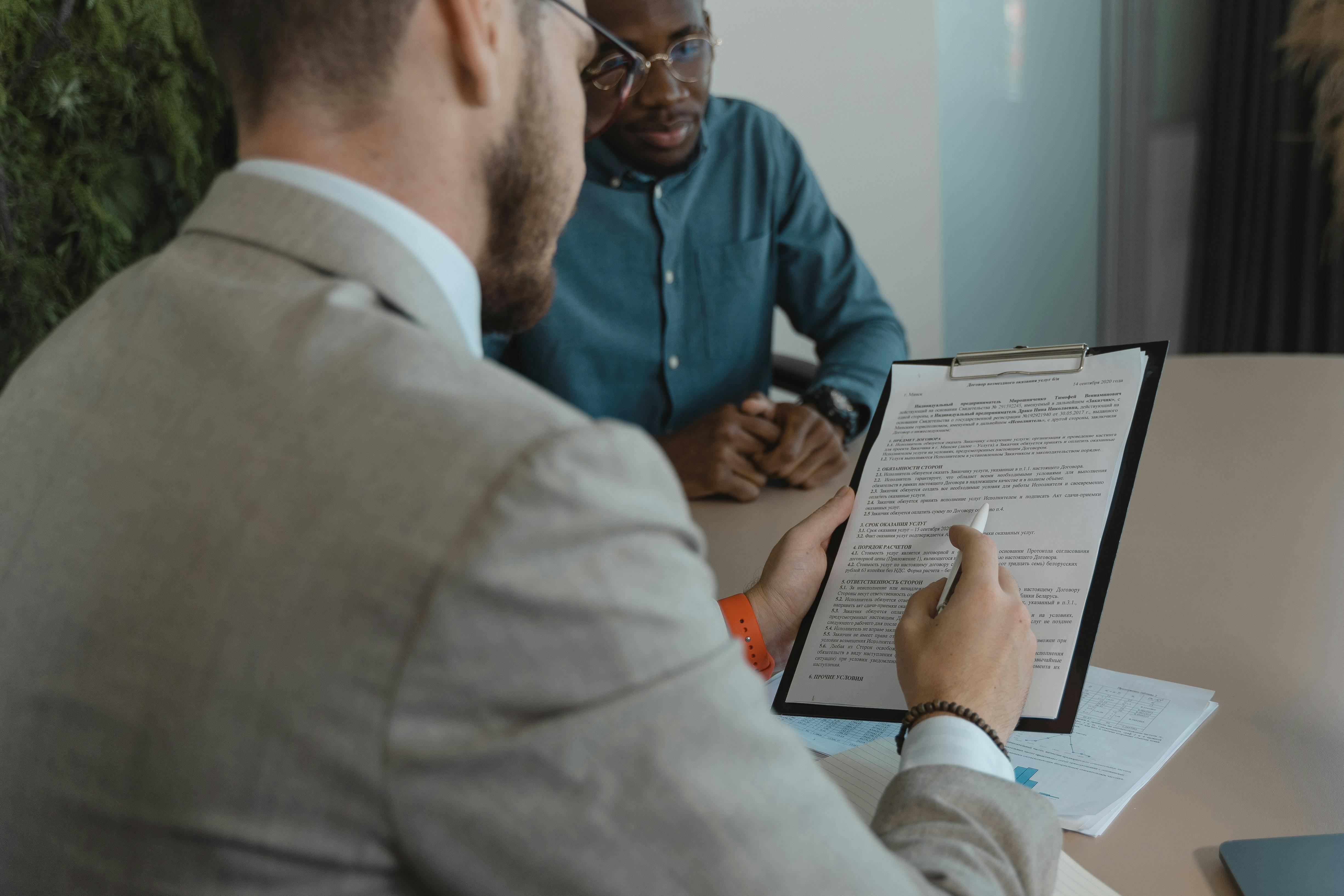 Two men in an office sit across a desk reviewing a document on a clipboard, with one pointing to the page while the other listens attentively; papers with charts lie on the table